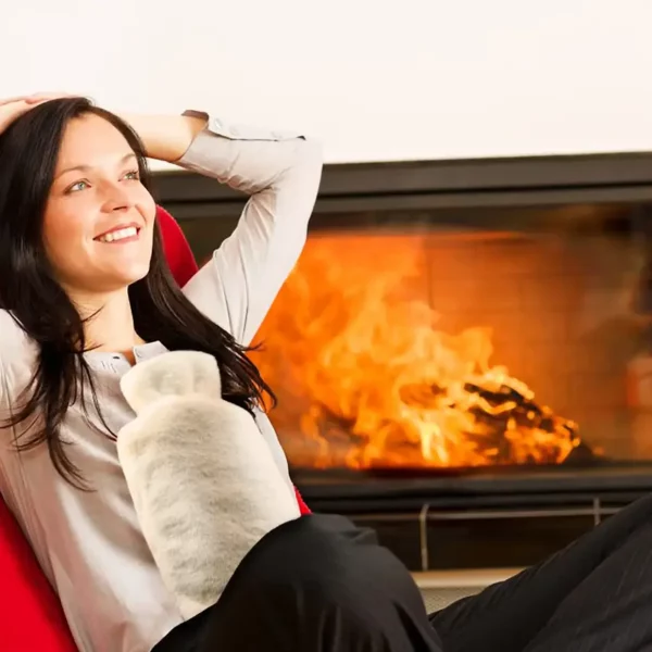 Une femme assise sur un fauteuil au coin du feu, avec une bouillotte Rissler sur le ventre